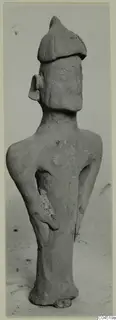 Terracotta figurine of a standing figure with a tall cap and bent arms, photographed against a plain grey background.