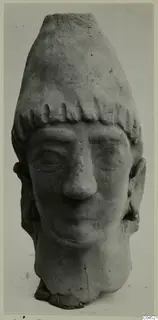 Terracotta head of a figure with a tall flat-topped headdress and fringe of curls, face with broad nose and downcast eyes.