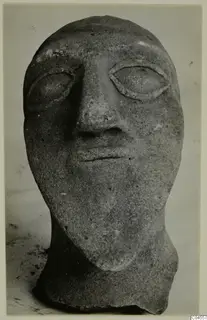 Front view of a grey stone head with almond-shaped eyes, a long nose and a thin mouth