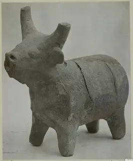 Terracotta figurine of a bull with upright horns and four short legs, photographed against a grey backdrop