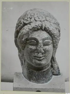 Limestone sculpture head from Arsos with curled hair, shown on a square stone base
