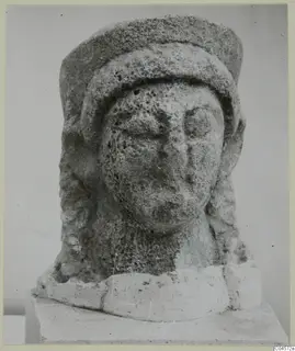 Limestone head sculpture from Arsos, with a tall cylindrical headdress and long locks of hair.