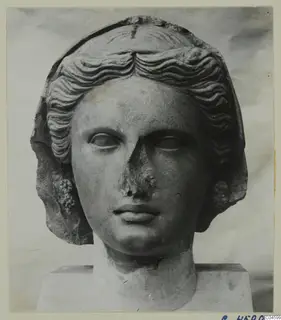 Stone head of a woman with wavy hair and a veil, nose damaged, set on a plinth