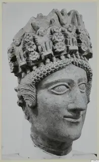 Carved limestone head of a figure wearing a tall headdress of rosettes and rounded motifs, photographed against a grey backdrop.