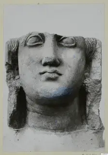 Stone statue head from Arsos, face frontal with large eyes, a broad nose and a small mouth, against a pale background.