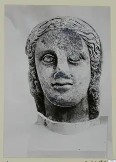 Stone statue head from Arsos, shown frontally on a block, with one eye partly closed and blue-grey staining on the face.