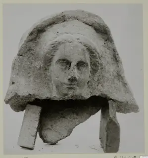 Carved stone head fragment with a hood-like headdress and worn face, propped on two rectangular blocks