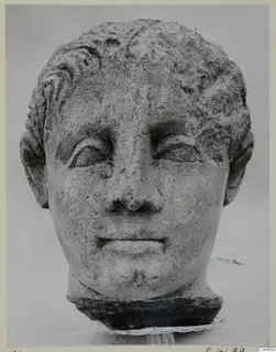 Carved stone head with short wavy hair and a worn face, set on a dark block against a pale background