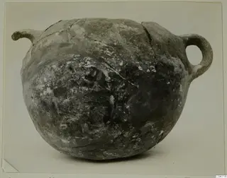 Round coarse-ware pot with a spout at left and a loop handle at right, shown against a plain background.