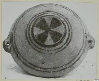 Pottery White Painted II: a flat, disc-shaped vessel with two pierced lugs and a dark six-spoked motif at the centre of its top