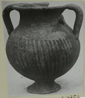 Black-slipped pottery jug with two loop handles, a wide body with vertical fluting, and a low foot, photographed against a pale background.