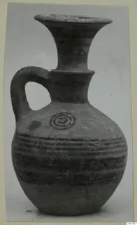 Pottery juglet with a trefoil-like flared neck, single loop handle, and dark painted bands and concentric circle on a buff body.