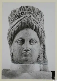 Stone statue head from Arsos, with a high headdress, wavy hair and carved rosettes, on a plinth.