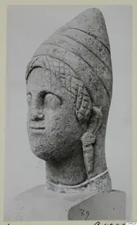 Stone statue head from Arsos with a tall cone-shaped headdress, shown in three-quarter view on a block base.