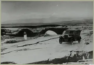 Volvo touring car on a cracked road, with an old stone arched bridge over a river behind it.