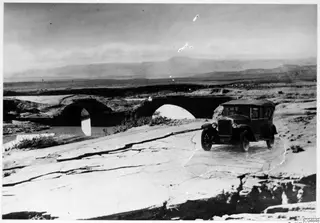 Dark boxy car on a flat, cracked stone road, with an arched stone bridge over a river behind it.