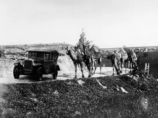 Early boxy car on a dirt road beside a line of camels, the lead camel carrying a rider, in a dry hilly landscape.