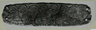 Long embossed metal strip with a row of profile faces and spiral motifs, edged with rows of small dots.