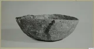 Side view of a thick-walled, rounded bowl with an uneven rim and a pitted, pale grey surface