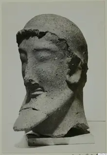 Sculpted head with a moustache and broken chin, from Mersinaki, shown on a wooden block