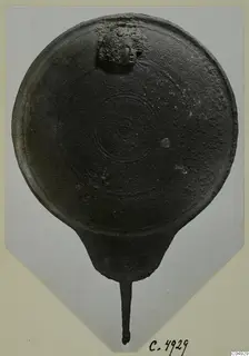 Round dark metal disc with a long tang and a small relief head of a figure with curly hair, from Marion