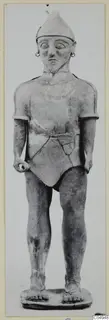 Standing terracotta figure in a conical cap, with ring earrings and a short belted garment, on a flat base.