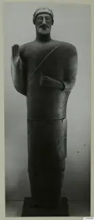 Standing statue from Mersinaki, one hand raised palm forward, bearded, with a cap-like head covering.