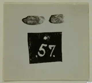 Two small worn objects above a black number label, shown on a light card mount