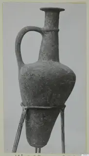 Base-ring ware juglet with a long neck, flared rim and loop handle, shown on a metal stand.