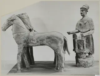 Terracotta group of a horse and a helmeted standing figure beside a rectangular frame, on a plain background.