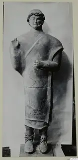 Standing bearded male terracotta statue on a plinth, photographed against a pale backdrop