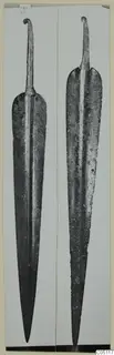 Two long leaf-shaped metal blades with curled tangs, photographed upright side by side.