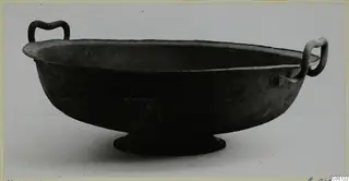 Dark metal bowl with two handles and a low ring foot, photographed on a pale background