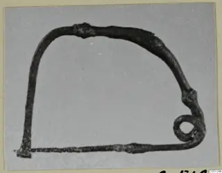 Dark, corroded metal wire object with a rounded top and a small coiled ring at one end, photographed on grey