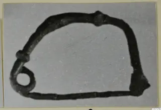 Iron object with a bent, rod-like form, a small ring at the lower left and a flattened end at the lower right, on a grey backdrop.