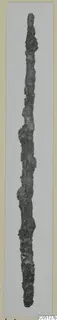 Long, thin corroded iron object photographed upright against a grey background, from the Kastros tombs at Lapithos