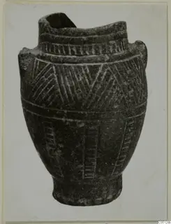 Dark stone vessel with incised hatched decoration and a broken rim, photographed on a light card