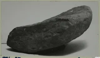 Stone tool from Petra tou Limniti, dark grey and elongated, photographed on a pale background