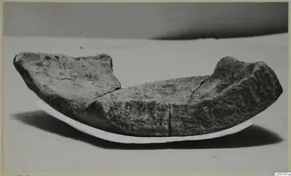 Curved stone object from Petra tou Limniti, broken along its upper edges, shown on a pale curved support.