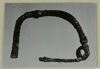 Corroded metal object bent into an open arch, with a small loop at the right end and a straight tail at the lower left