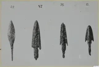 Four corroded bronze weapon points from Ayios Iakovos laid in a row, each with a handwritten number label above it.