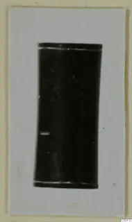 Dark cylindrical find with pale rims at top and bottom, mounted on a light card from the Ayios Iakovos Bronze Age finds
