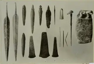 Bronze weapons and tools from Lapithos Tomb 315 B-C laid out on a grey background, with a rough object at the right