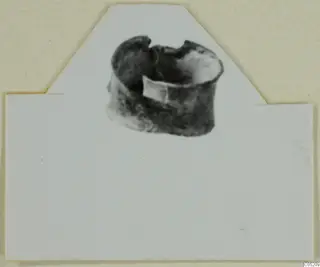 Small dark, corroded ring-like object from Enkomi Tomb 3, shown as a print on a cut-out white card mount