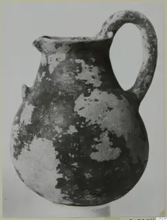 Black Slip Ware jug with a loop handle and a small spout, its dark surface flaking to lighter patches