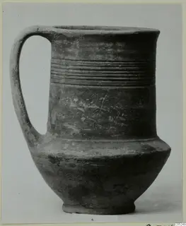 Handled pottery vessel with a tall body, a band of horizontal grooves and a flared rim, dark grey-brown surface