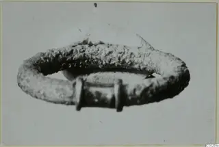 Ring of corroded metal with a flat raised oval plate on top and a rectangular fitting at the front