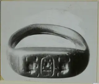 Metal finger ring with a wide oval bezel bearing an engraved design, shown on a pale photo mount.
