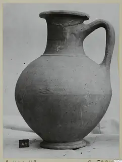 Plain pottery jug with a single handle and a flared rim, shown on a white cloth against a pale backdrop.