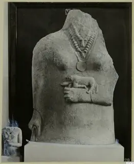 Headless limestone statue torso with a beaded necklace, a disc pendant, and a small carved animal in relief across the belly.